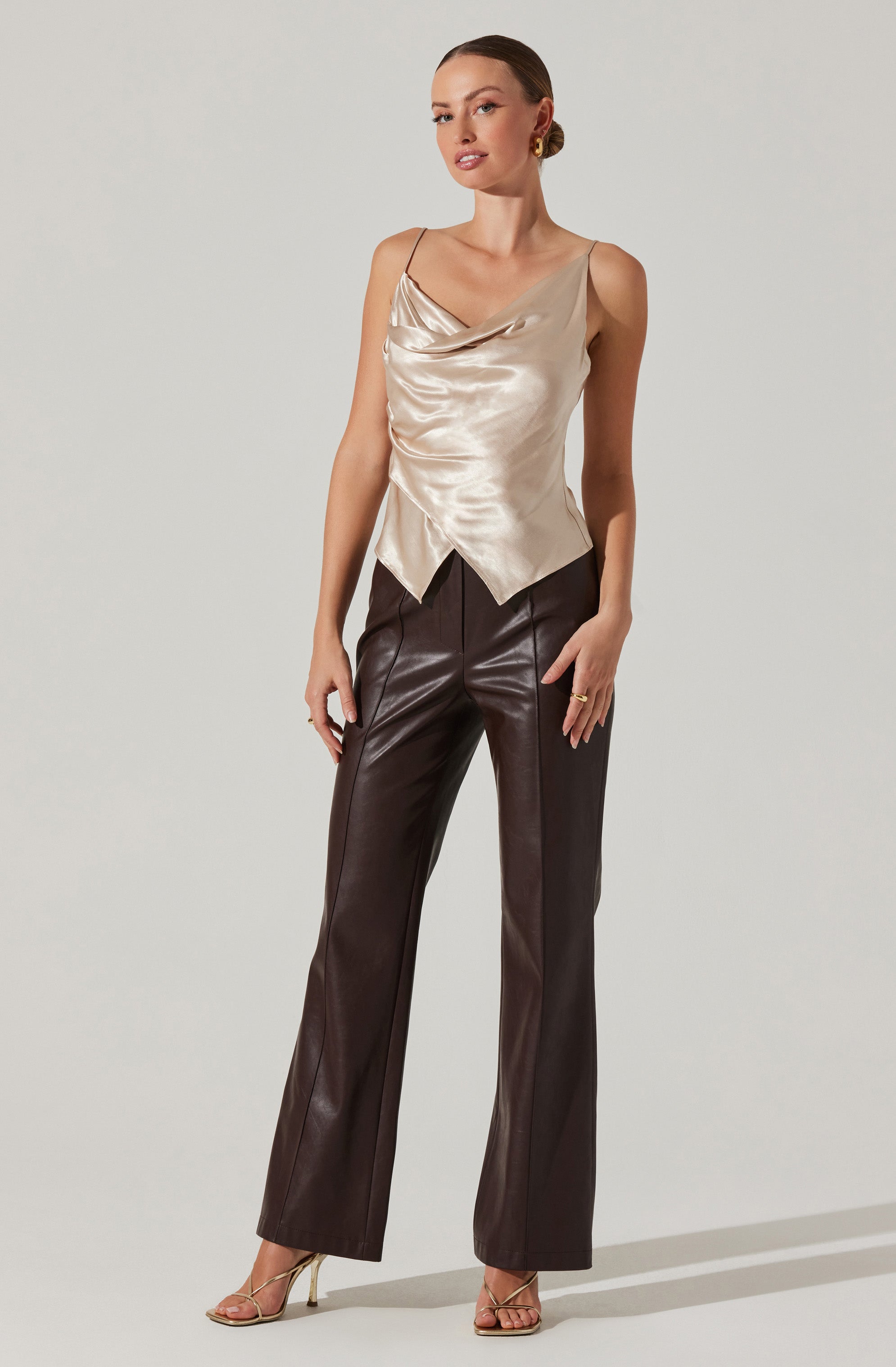 A woman is wearing a sleeveless, satin-like champagne-colored top with a draped cowl neckline and a pointed hem. She pairs it with high-waisted, straight-leg dark brown leather pants. She completes the look with beige strappy high-heeled sandals.