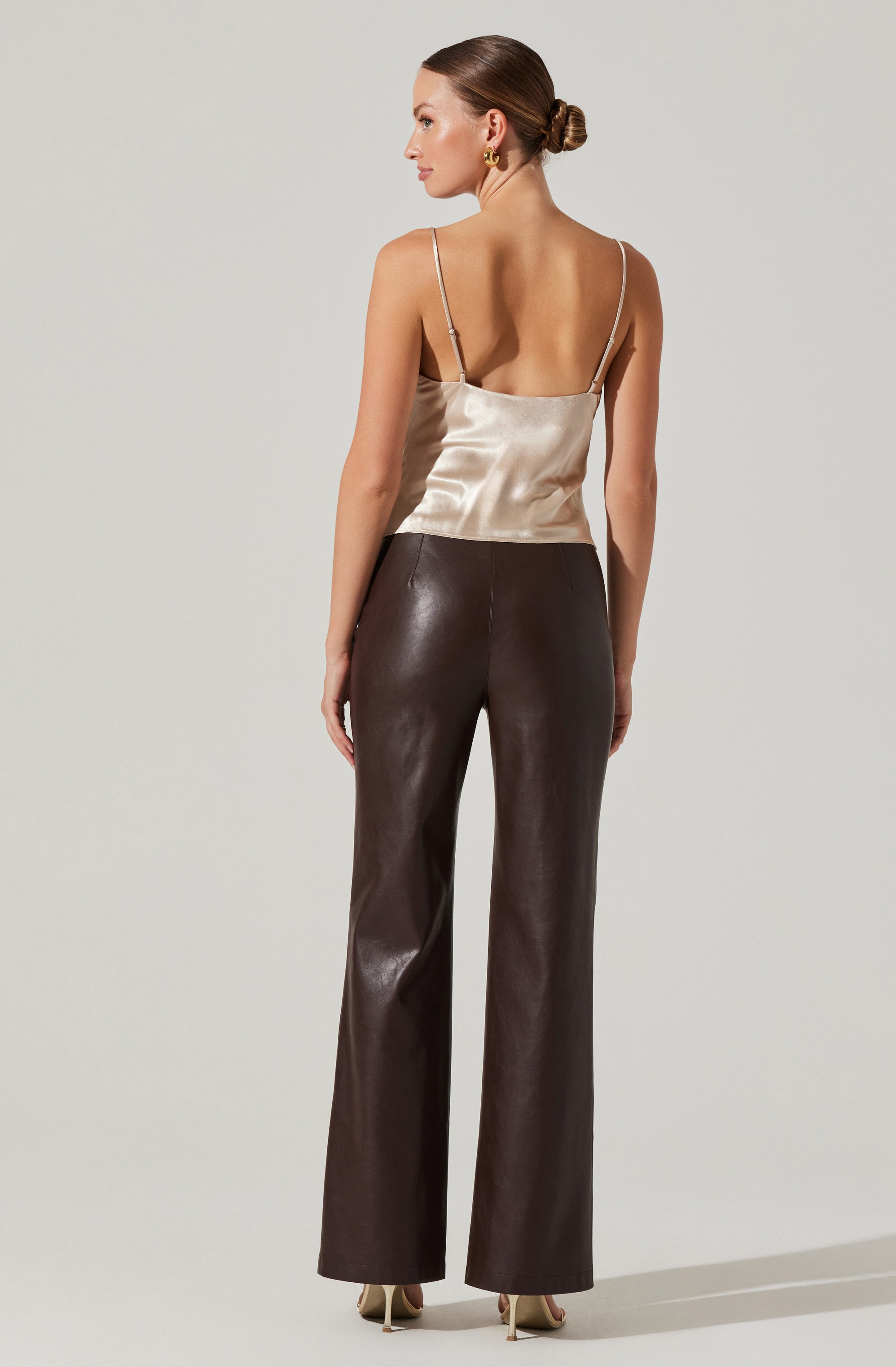 A woman is wearing a silky, beige spaghetti-strap top with a fitted waist and wide-leg, dark brown leather pants. She has her hair styled in a low bun and is wearing gold hoop earrings.