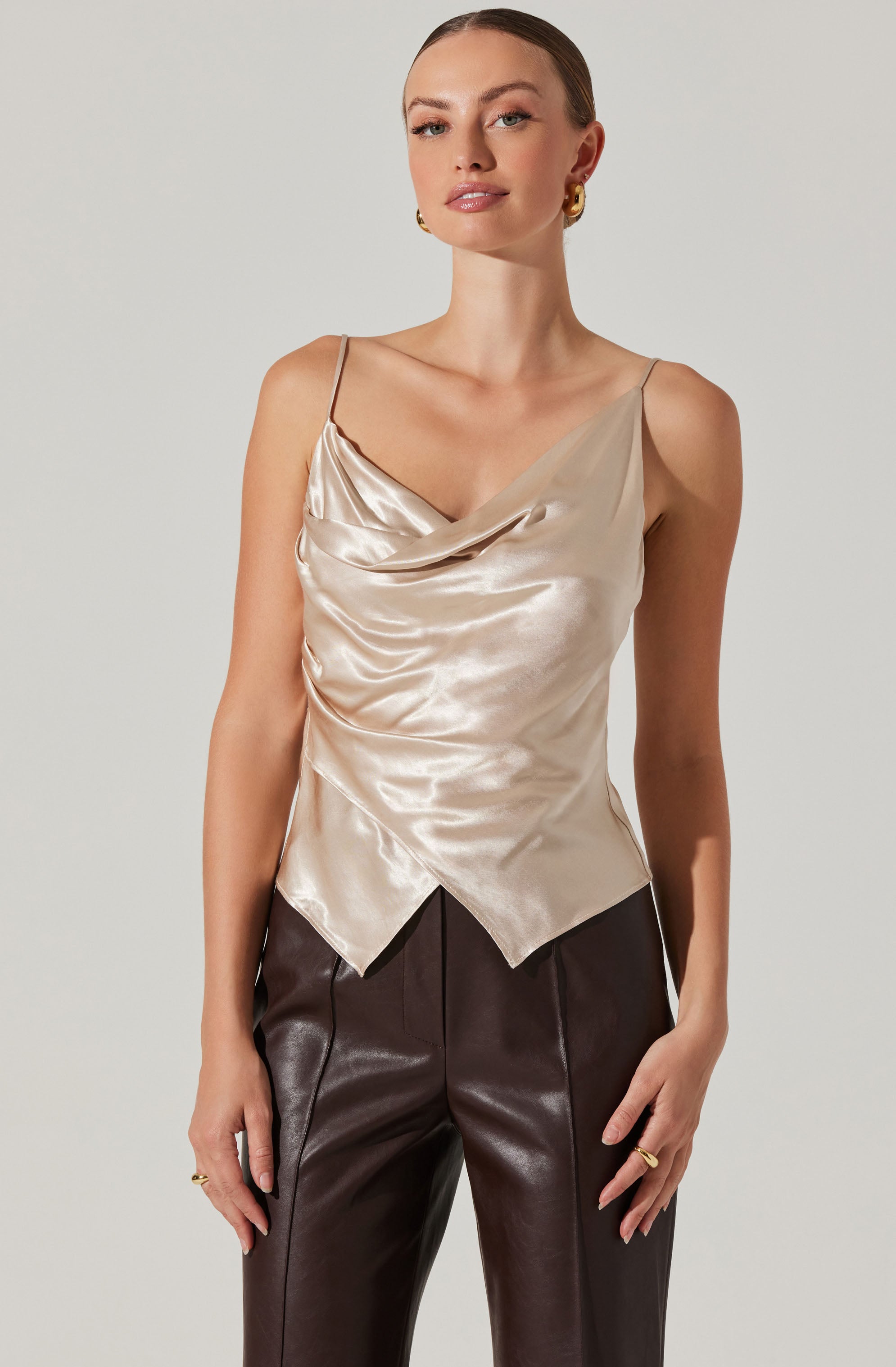 The woman is wearing a shiny, cream-colored satin camisole with a draped neckline and a pointed hemline. She pairs it with high-waisted, dark brown leather pants that have visible seams running down the front.