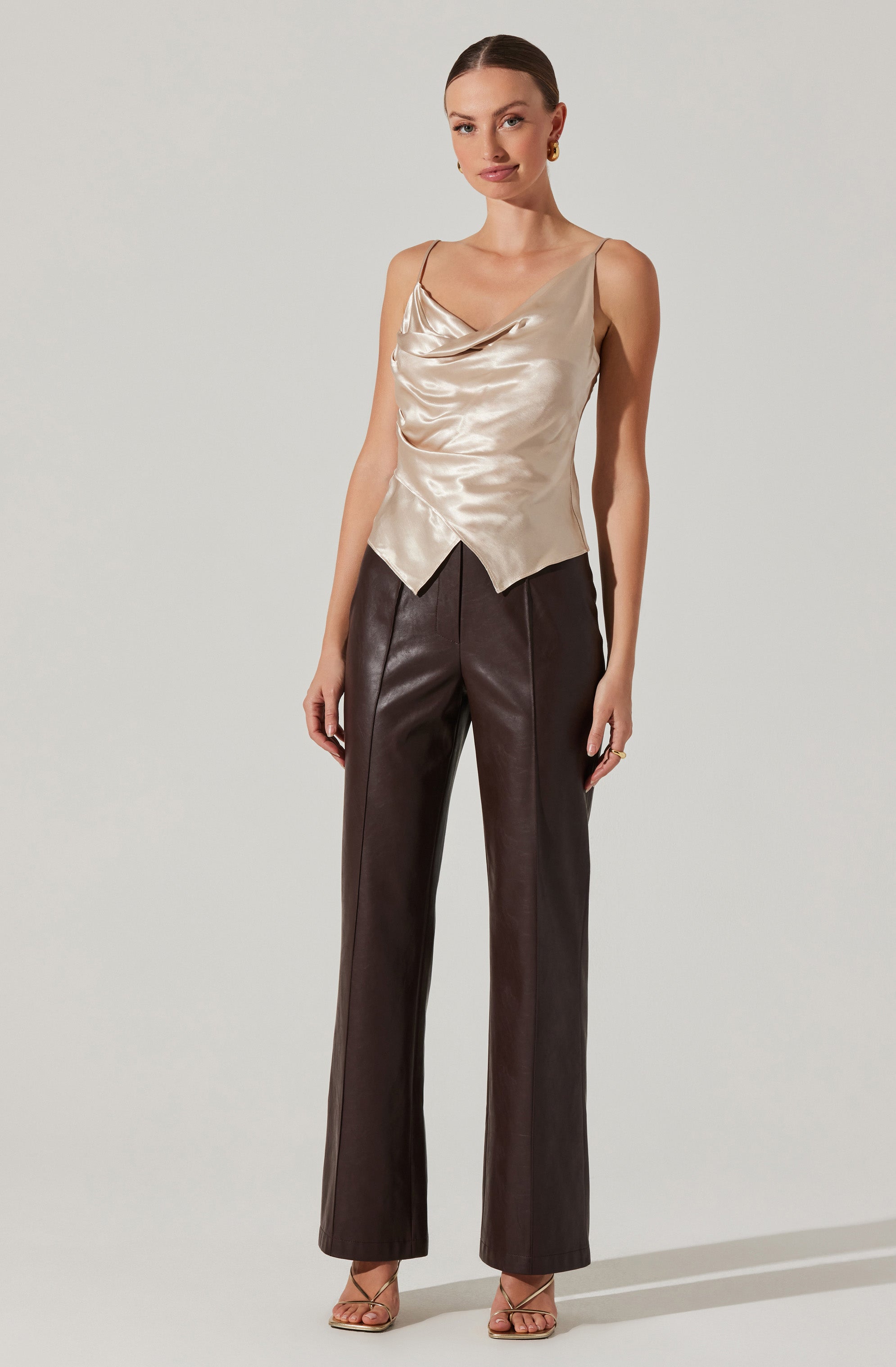 A woman is wearing a sleeveless, satin champagne-colored top with a draped neckline and a pointed hemline. She is also dressed in high-waisted, straight-leg brown leather pants. She accessorized with gold earrings and rings, and she is wearing beige open-toe high-heeled sandals.