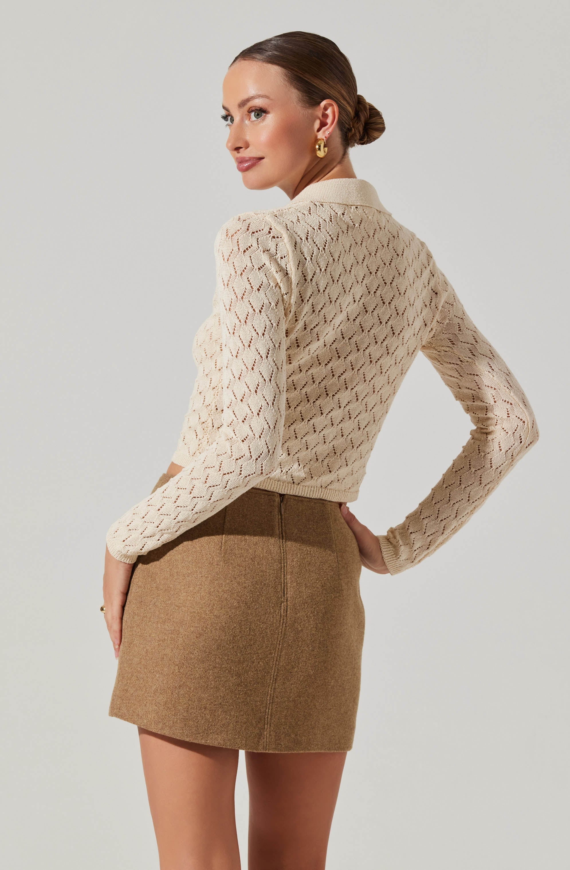 A woman is wearing a cream-colored, long-sleeved, open-knit sweater with a diamond pattern, paired with a high-waisted, camel-colored, woolen mini skirt.