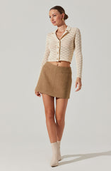 A woman is wearing a cream-colored, long-sleeve, button-up cardigan with a textured, open-knit pattern. She pairs it with a high-waisted, A-line mini skirt in a light brown/tan color. Her footwear consists of ankle-high, beige boots. She has her hair styled in a sleek, low bun and is accessorized with gold earrings.