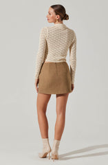 A woman is wearing a cream-colored, long-sleeved, cable-knit sweater with a subtle diamond pattern. She pairs it with a high-waisted, camel-colored, A-line mini skirt made of a wool or wool-blend fabric. She completes the outfit with shiny, beige ankle boots with high heels.