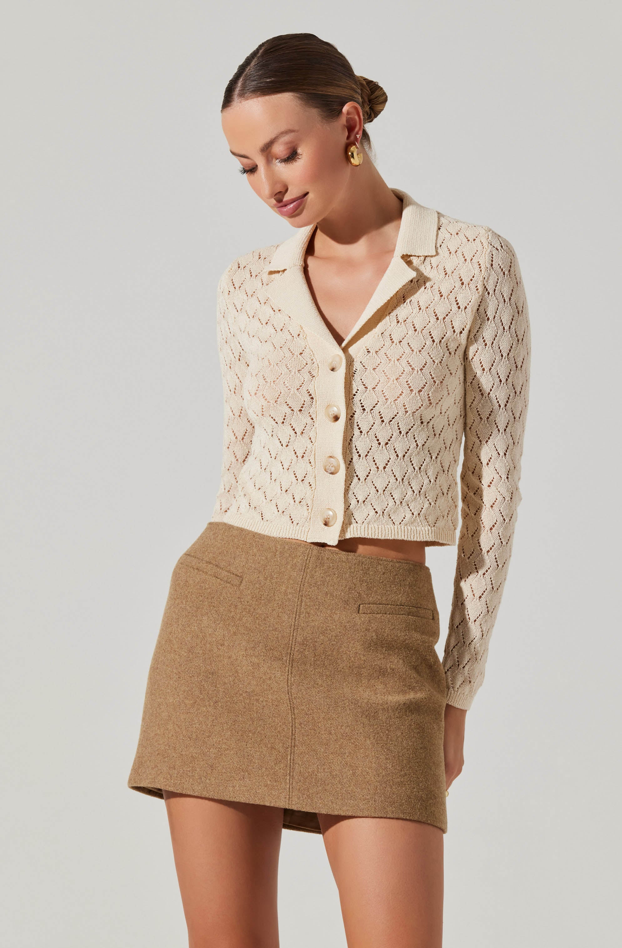 A woman is wearing a cream-colored, textured knit cardigan with a diamond pattern, featuring large buttons down the front and a collar. She pairs it with a high-waisted, camel-colored tweed mini skirt with a straight cut and two small back pockets.