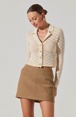 A woman is wearing a cream-colored, textured knit cardigan with a diamond pattern, featuring large buttons down the front and a collar. She pairs it with a high-waisted, camel-colored tweed mini skirt with a straight cut and two small back pockets.