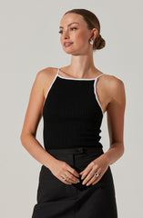 A woman is wearing a black ribbed tank top with white trim along the neckline and straps. She has her hair styled in a low bun and is accessorized with earrings and rings. She is also wearing black pants.