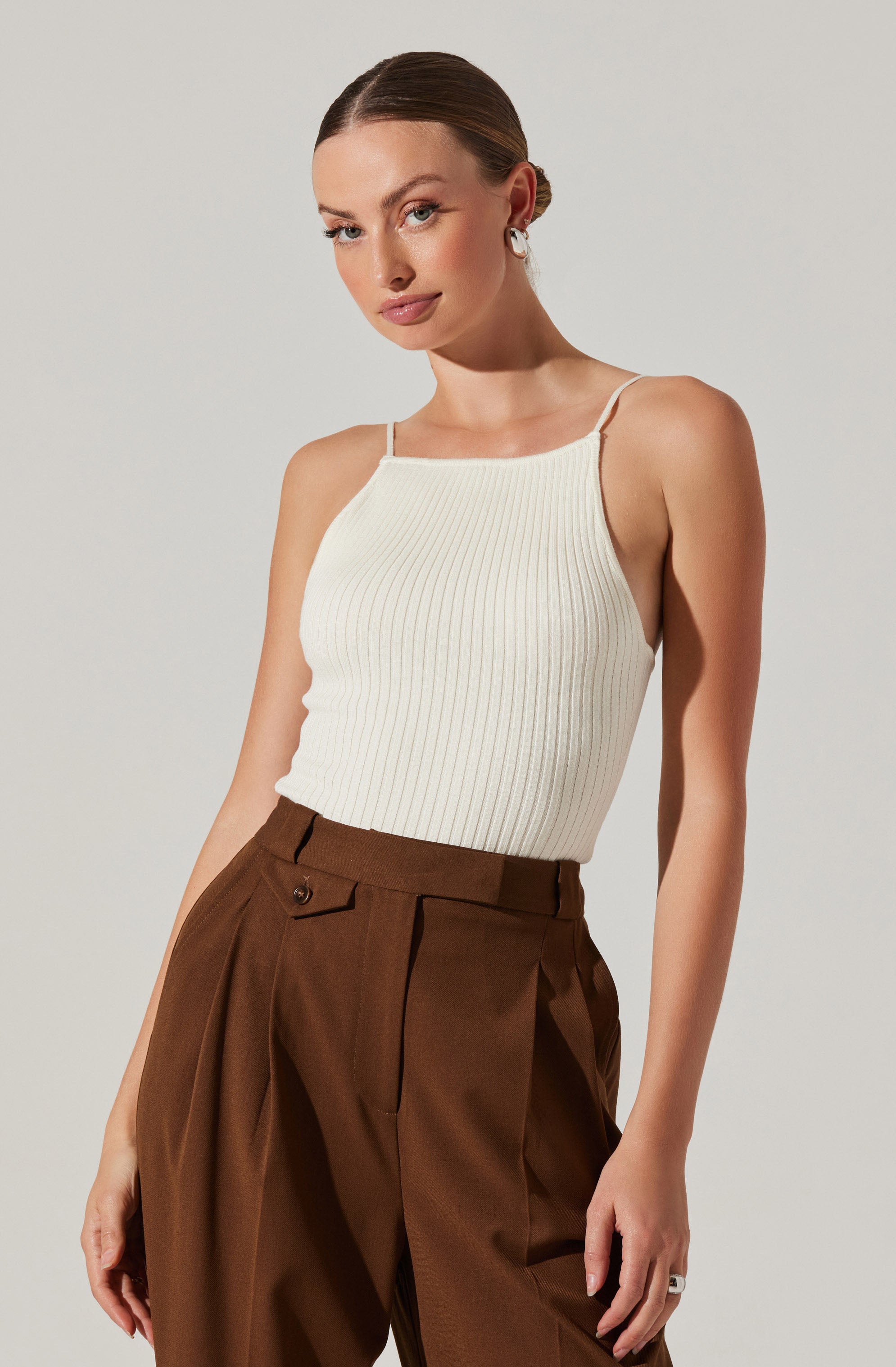 A woman is wearing a sleeveless, ribbed, cream-colored top with thin spaghetti straps. She pairs it with high-waisted, brown, pleated trousers that have a wide-leg fit and feature a belt loop, pockets, and a button detail at the waist.