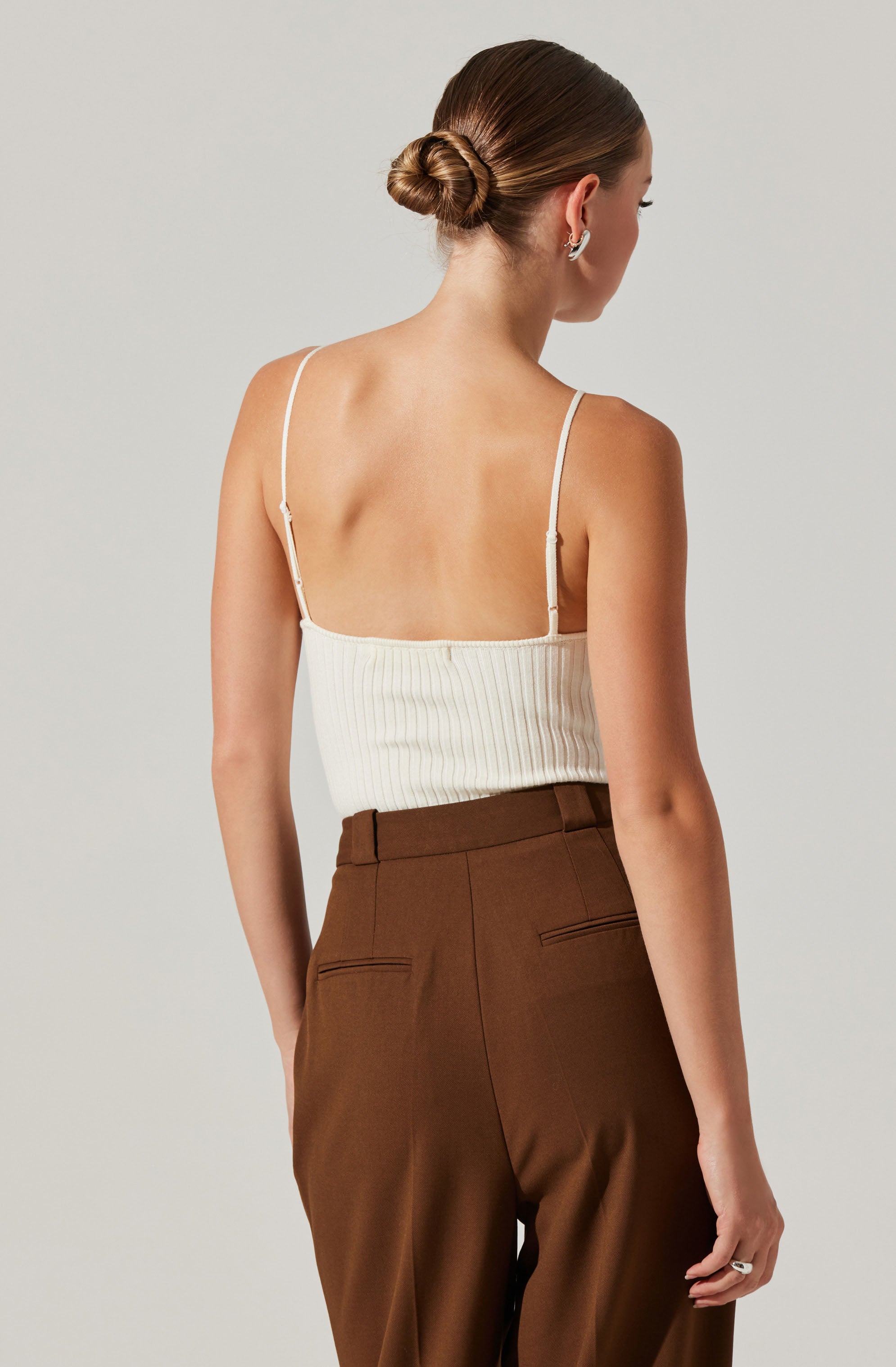 A woman wearing a white ribbed tank top with thin spaghetti straps and brown high-waisted trousers. The tank top is fitted and ends just above her hips. The trousers have a tailored fit with two welt pockets on the back.
