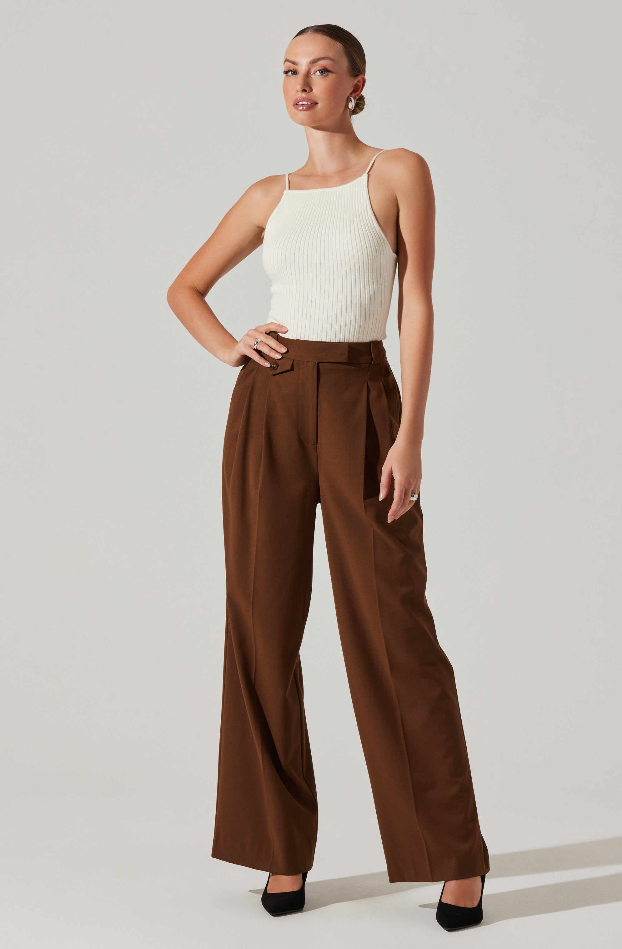 A woman wearing a sleeveless, ribbed, white tank top with spaghetti straps and high-waisted, wide-leg brown trousers. She has her left hand on her hip and is wearing black high heels.