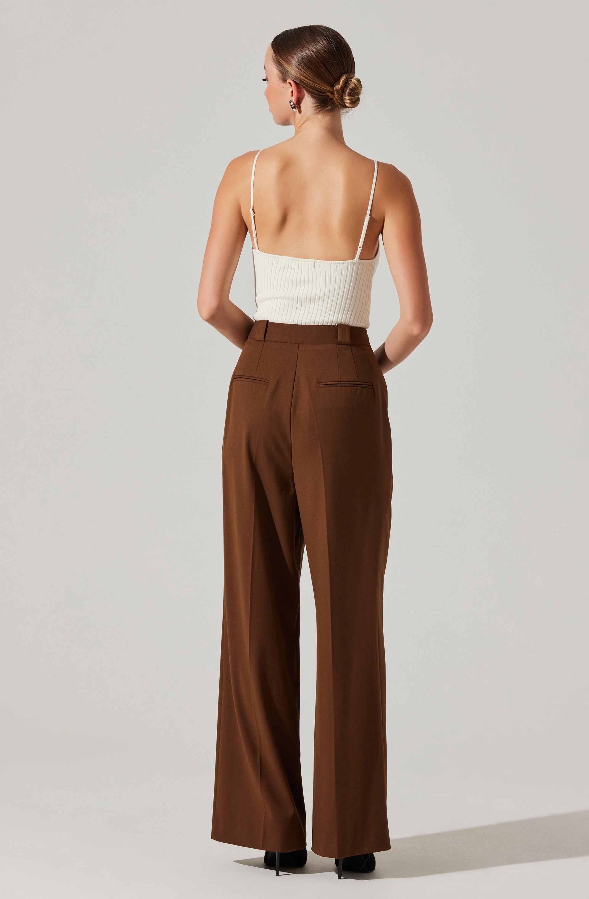 A woman is wearing a sleeveless, cream-colored ribbed top with thin spaghetti straps. She is also wearing high-waisted, wide-leg brown trousers with back pockets.