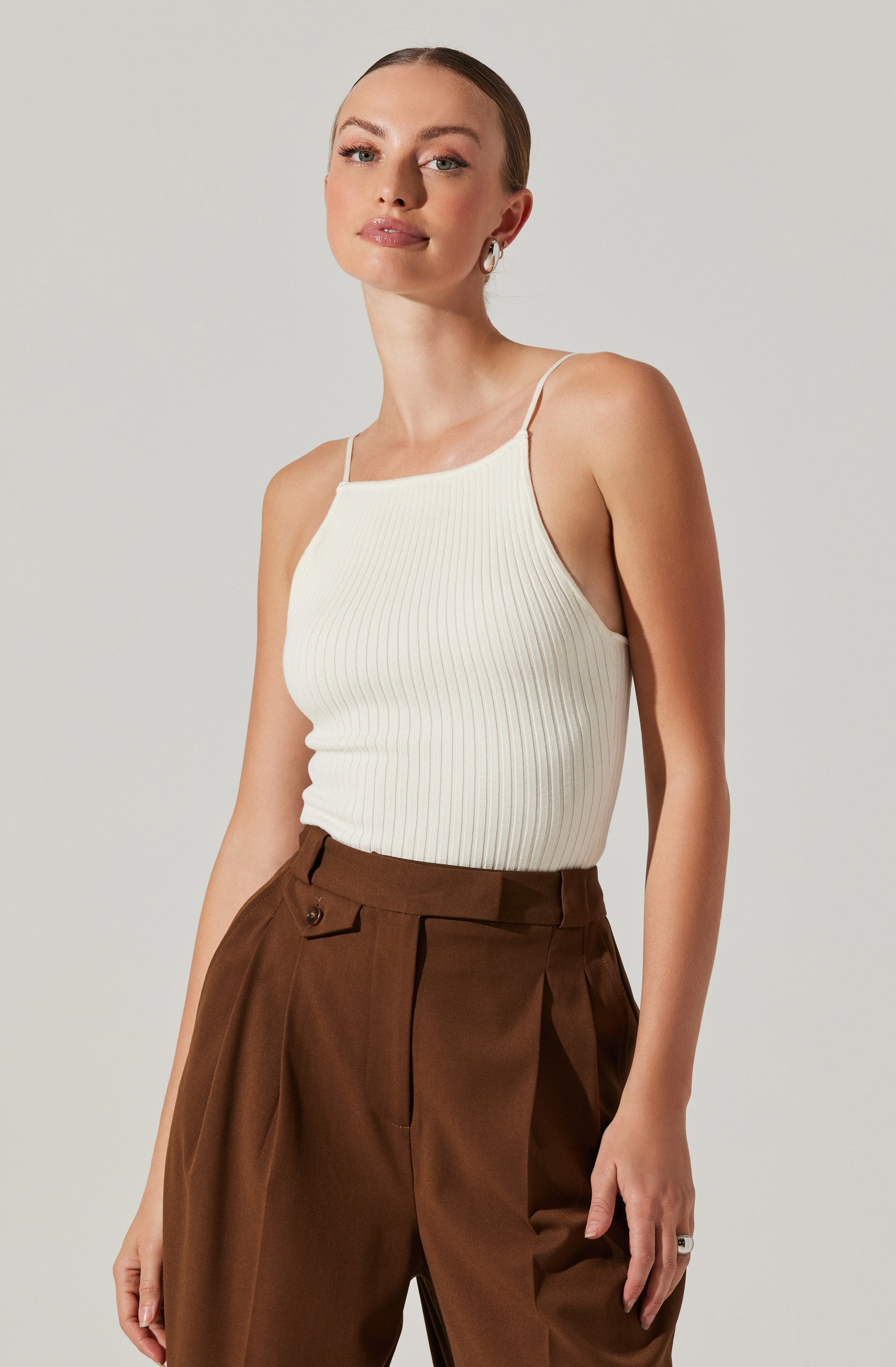 A woman is wearing a white, ribbed, spaghetti-strap tank top tucked into high-waisted brown trousers with pleats and a button detail at the waistband.