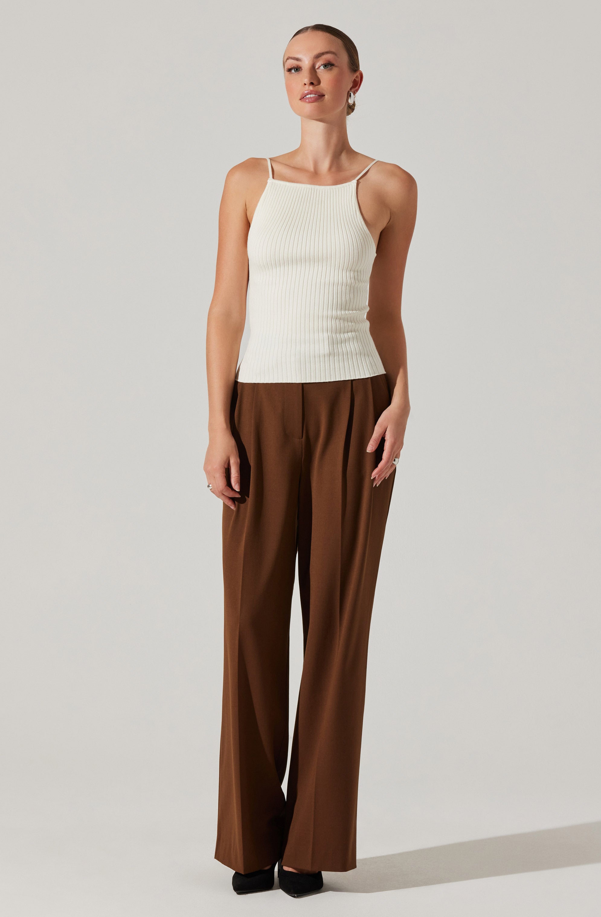 A woman is wearing a white, ribbed, sleeveless top with thin spaghetti straps. She pairs it with high-waisted, wide-leg brown trousers. She is also wearing black shoes and a single earring.