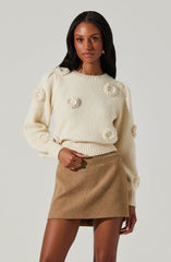 A woman is wearing a cream-colored, fuzzy sweater with textured, fluffy circular embellishments scattered across the front and sleeves. She is also wearing a high-waisted, camel-colored, A-line mini skirt made of a soft, wool-like material.