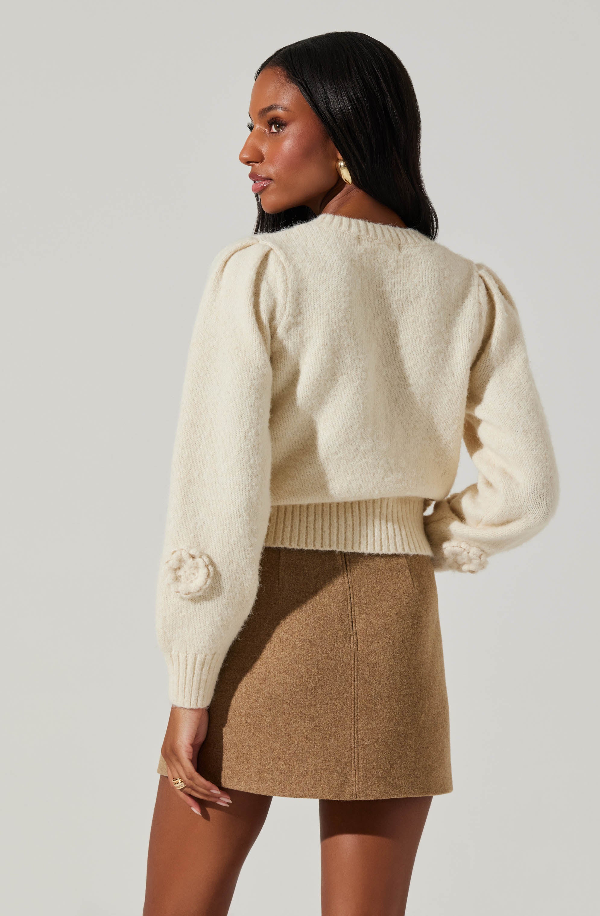 A woman is wearing a cream-colored, long-sleeved knit sweater with subtle puffed shoulders and decorative pom-poms on the sleeves. She pairs it with a high-waisted, camel-colored mini skirt made of a textured fabric.