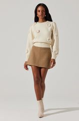 A woman is wearing a cream-colored, long-sleeved sweater with textured, circular details and a ribbed hem. She pairs it with a high-waisted, camel-colored, A-line mini skirt and matching cream-colored ankle boots.