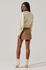 A woman is wearing a cream-colored, long-sleeved, fuzzy sweater with a slightly loose fit and ribbed cuffs. She pairs it with a high-waisted, short brown skirt made of a textured, wool-like fabric. She completes her outfit with knee-high, beige patent leather high-heeled boots.
