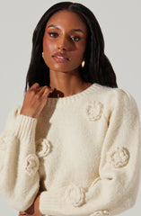 A woman wearing a cream-colored, knitted sweater with textured, fluffy floral appliqués on the front and sleeves. The sweater has a round neckline and ribbed cuffs. She is also wearing large, gold hoop earrings.