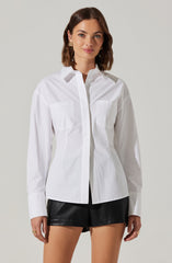 A woman is wearing a long-sleeved white button-up shirt with two chest pockets. The shirt has a classic collar and a slightly fitted silhouette. She pairs the shirt with black leather shorts.