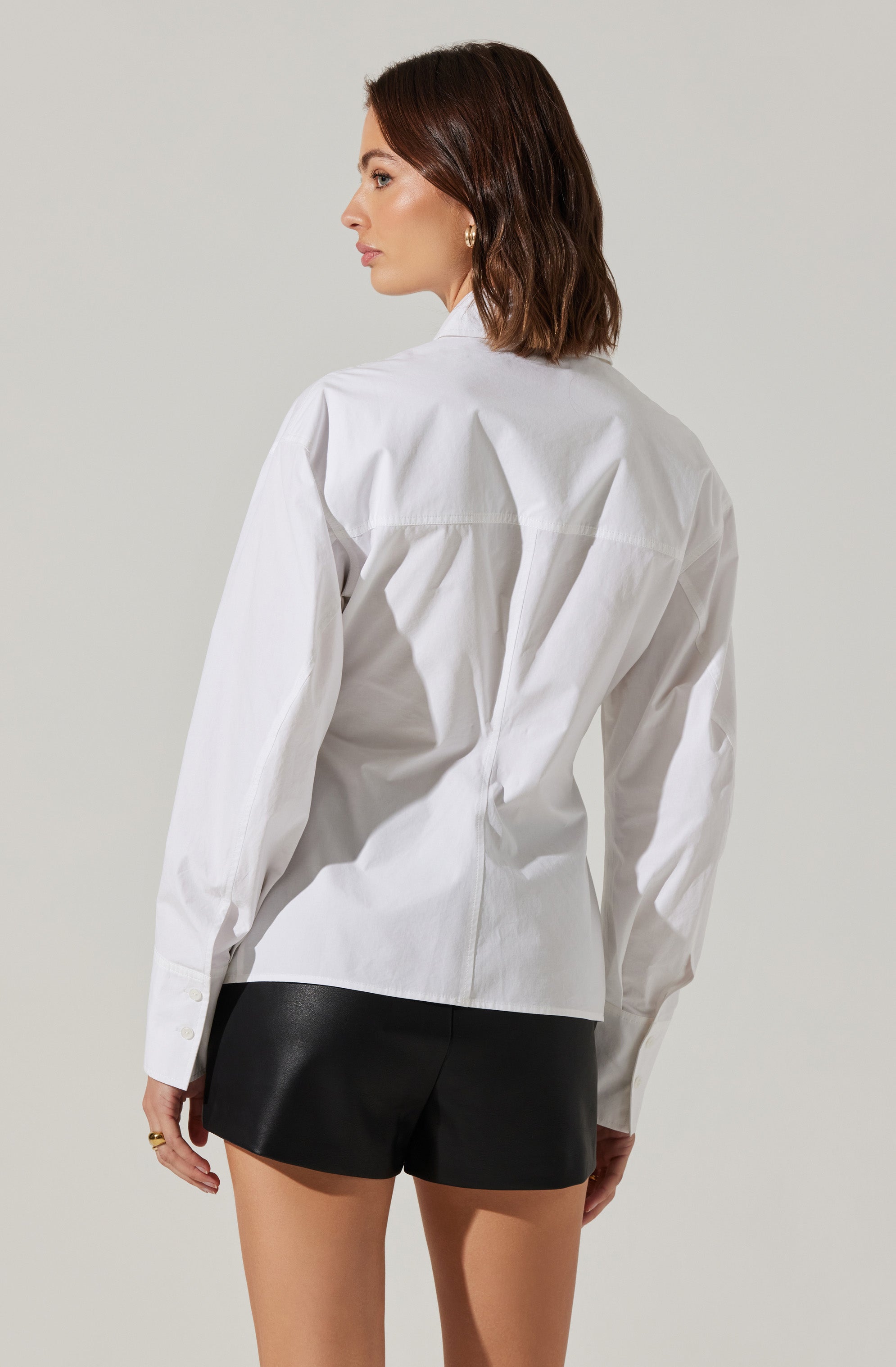 The image shows a woman wearing a white button-up shirt with long sleeves, featuring a classic collar and a tailored fit. She is also wearing black shorts. The shirt has a slightly loose fit with some subtle pleats and is made of a crisp fabric.