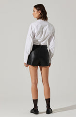 A woman is wearing a loose-fitting white button-up shirt with long sleeves, paired with high-waisted black leather shorts. She has on sheer black ankle socks and shiny black loafers.