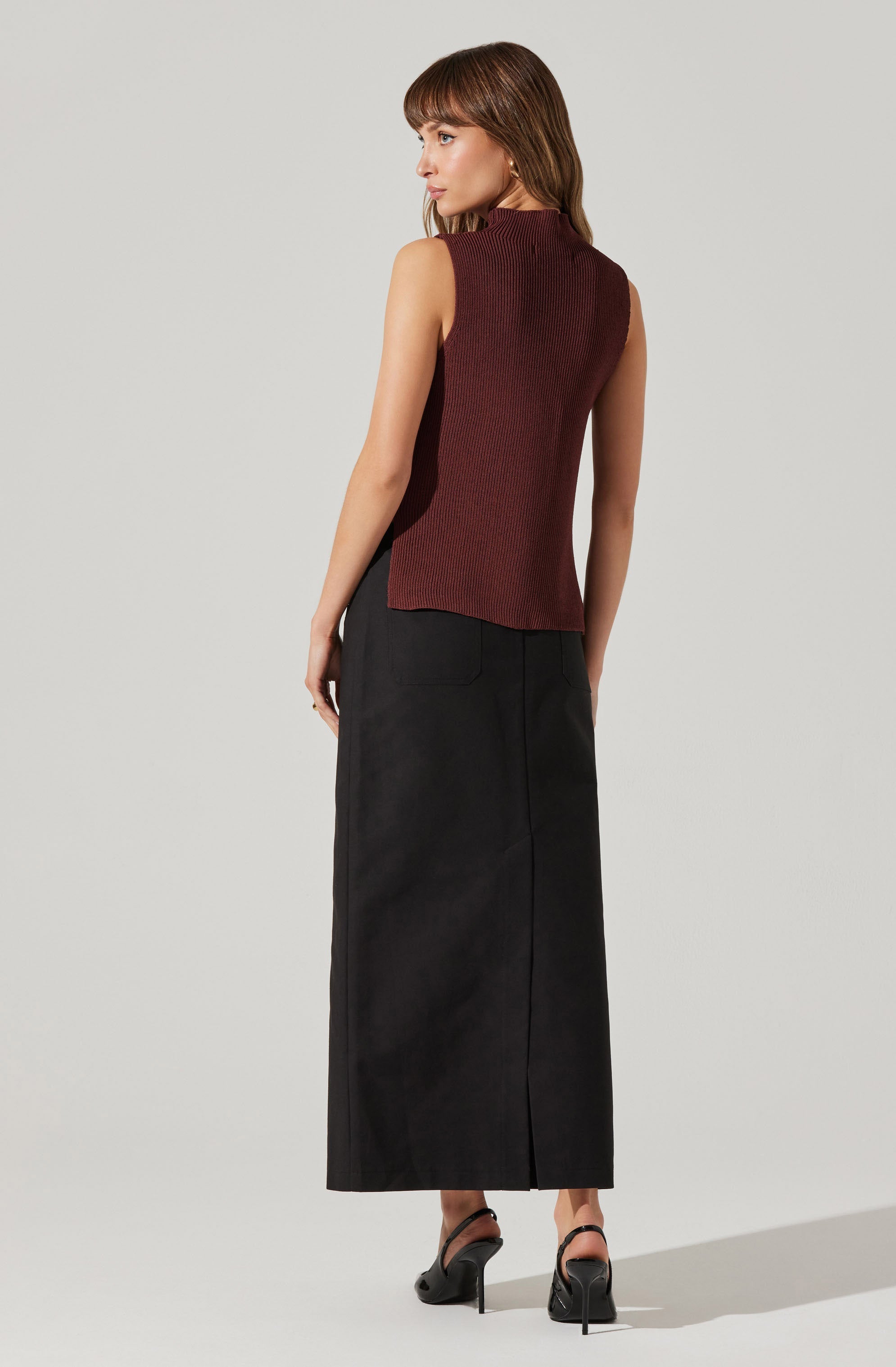 A woman is wearing a sleeveless, ribbed, high-neck top in a dark burgundy color and a long, black, ankle-length skirt. She is also wearing black high-heeled sandals with ankle straps.