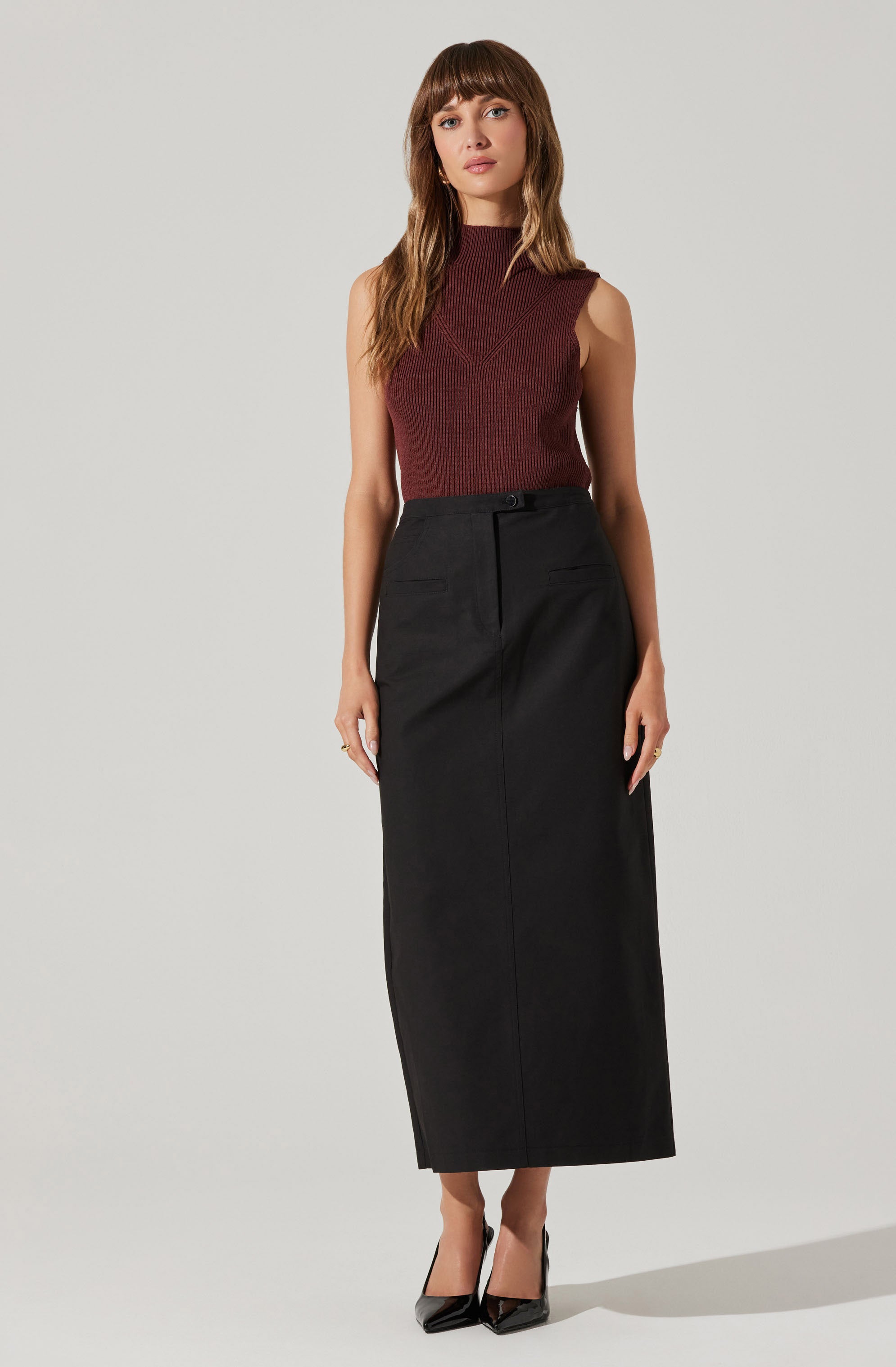 A woman is wearing a sleeveless, high-neck maroon knit top paired with a long, black high-waisted skirt that reaches her ankles. She is also wearing black pointed-toe high heels.