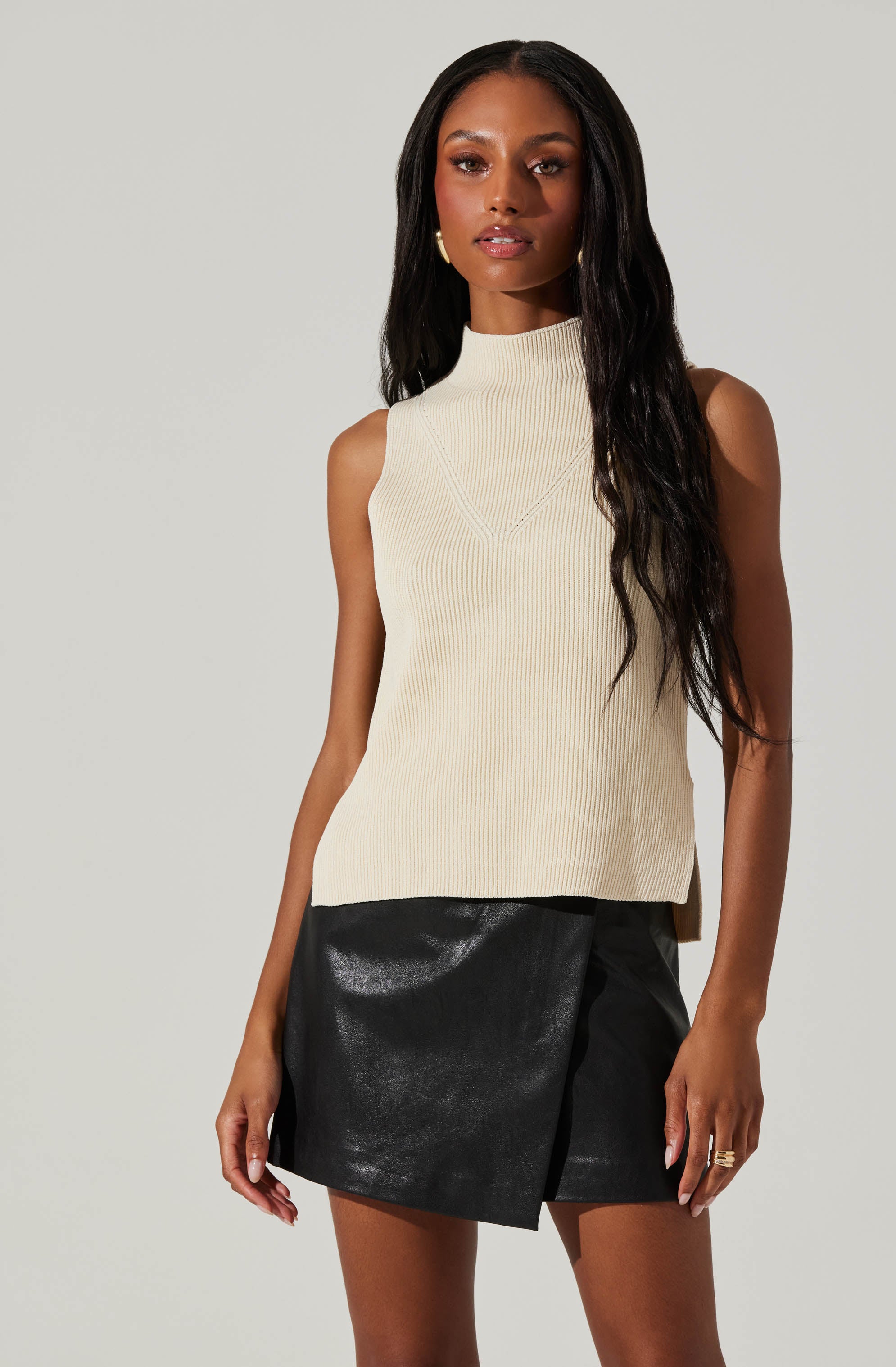 A woman is wearing a sleeveless, high-neck, cream-colored ribbed knit top paired with a black leather mini skirt that features a wrap style with a slight slit.