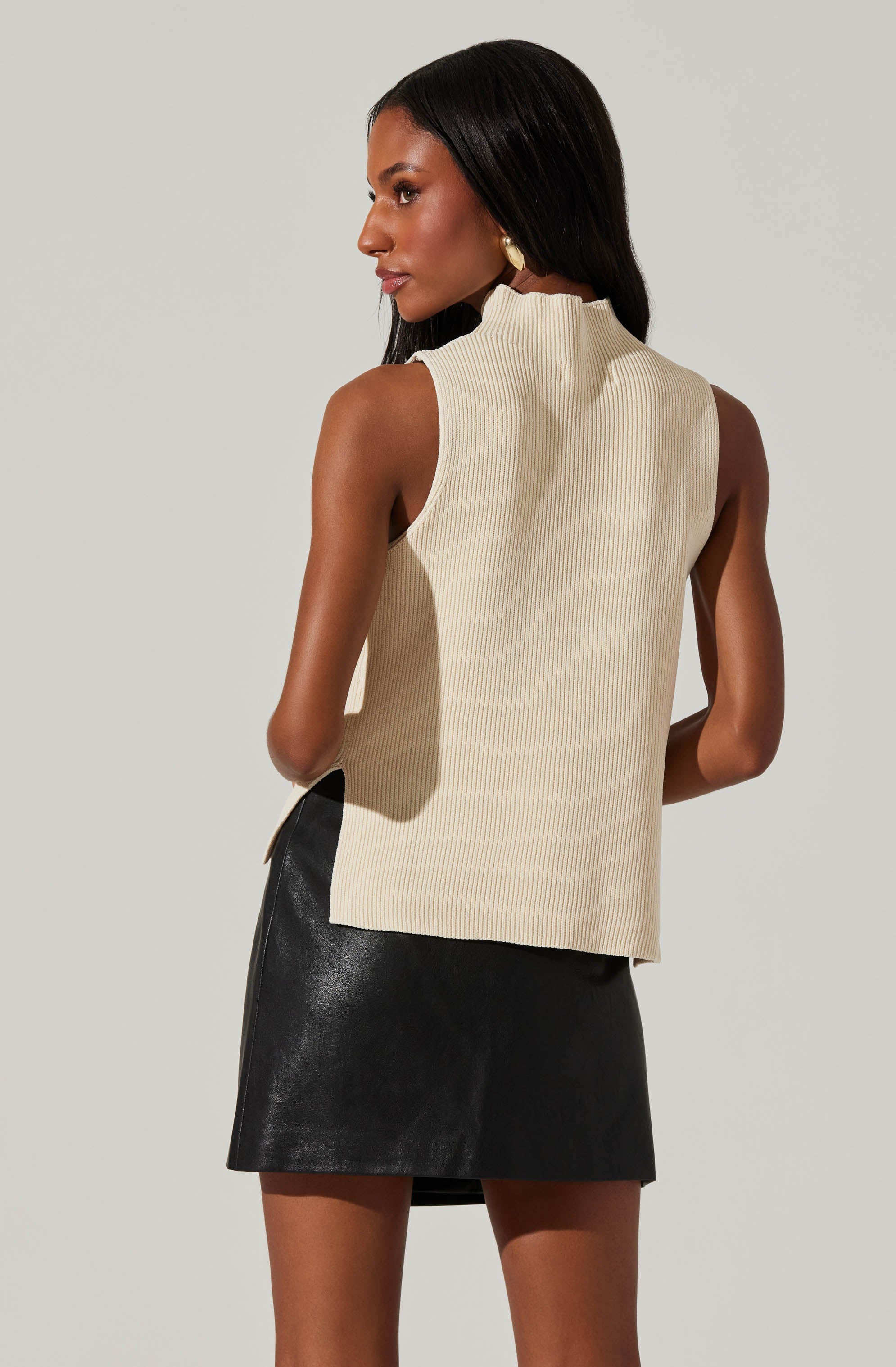 A woman is wearing a sleeveless, high neckline, cream-colored ribbed top paired with a black leather mini skirt.