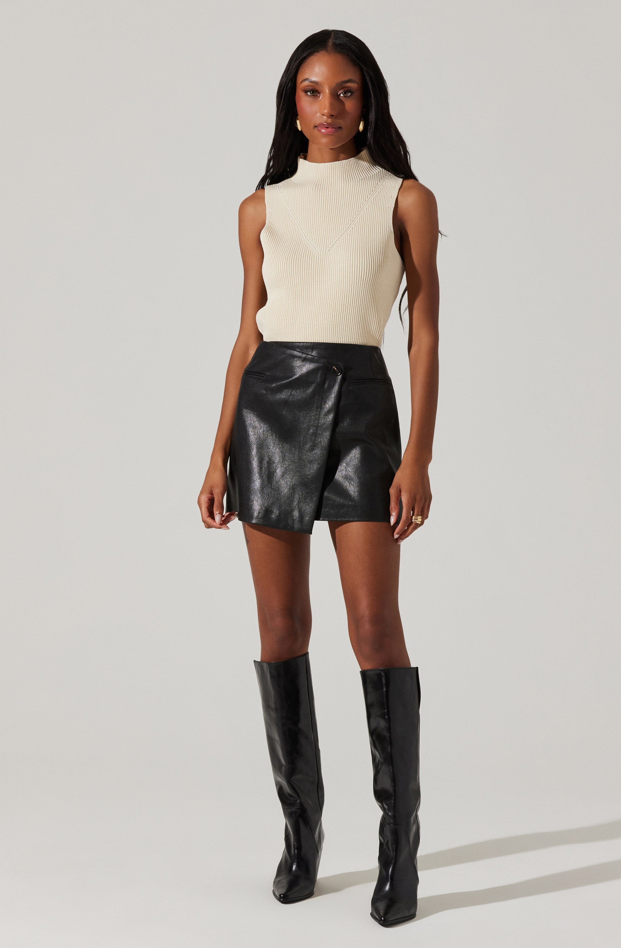 A woman is wearing a sleeveless, high-neck, cream-colored knit top paired with black leather shorts that have a zipper detail on the side. She is also wearing tall, shiny black leather knee-high boots.