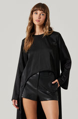 A woman is wearing a black long-sleeve top with a loose fit and subtle pleats on the front, paired with high-waisted black leather shorts.