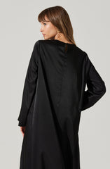 A woman is wearing a long, black, satin-like dress or coat with a high neckline and long sleeves. The back of the garment features a small keyhole opening at the neck, fastened with a button. The fabric has a smooth and slightly shiny texture.