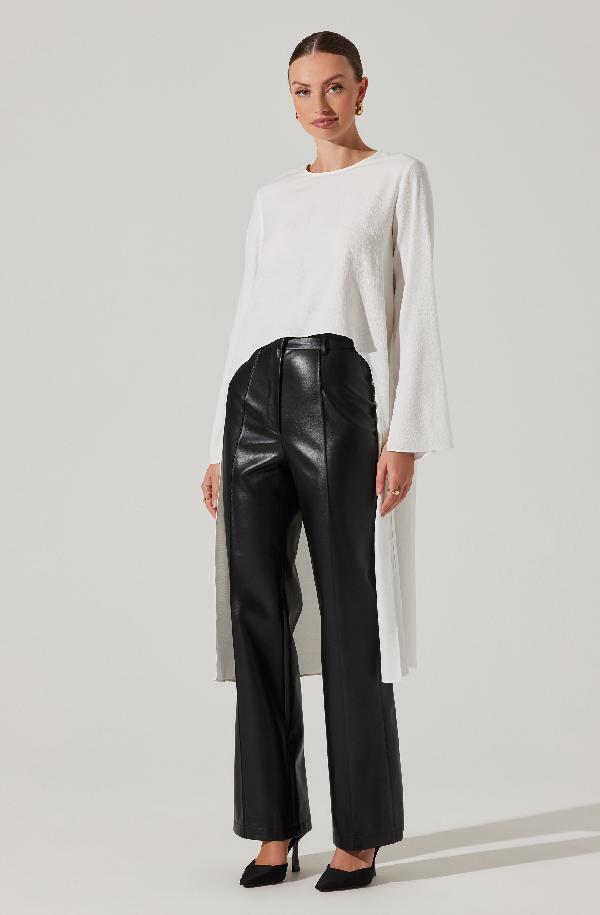 A woman is wearing a long-sleeved, white, textured top with a round neckline that extends to below her knees. She has paired it with high-waisted, black leather wide-leg trousers featuring vertical seam detailing along the front. She is also wearing black high-heeled shoes and gold hoop earrings.