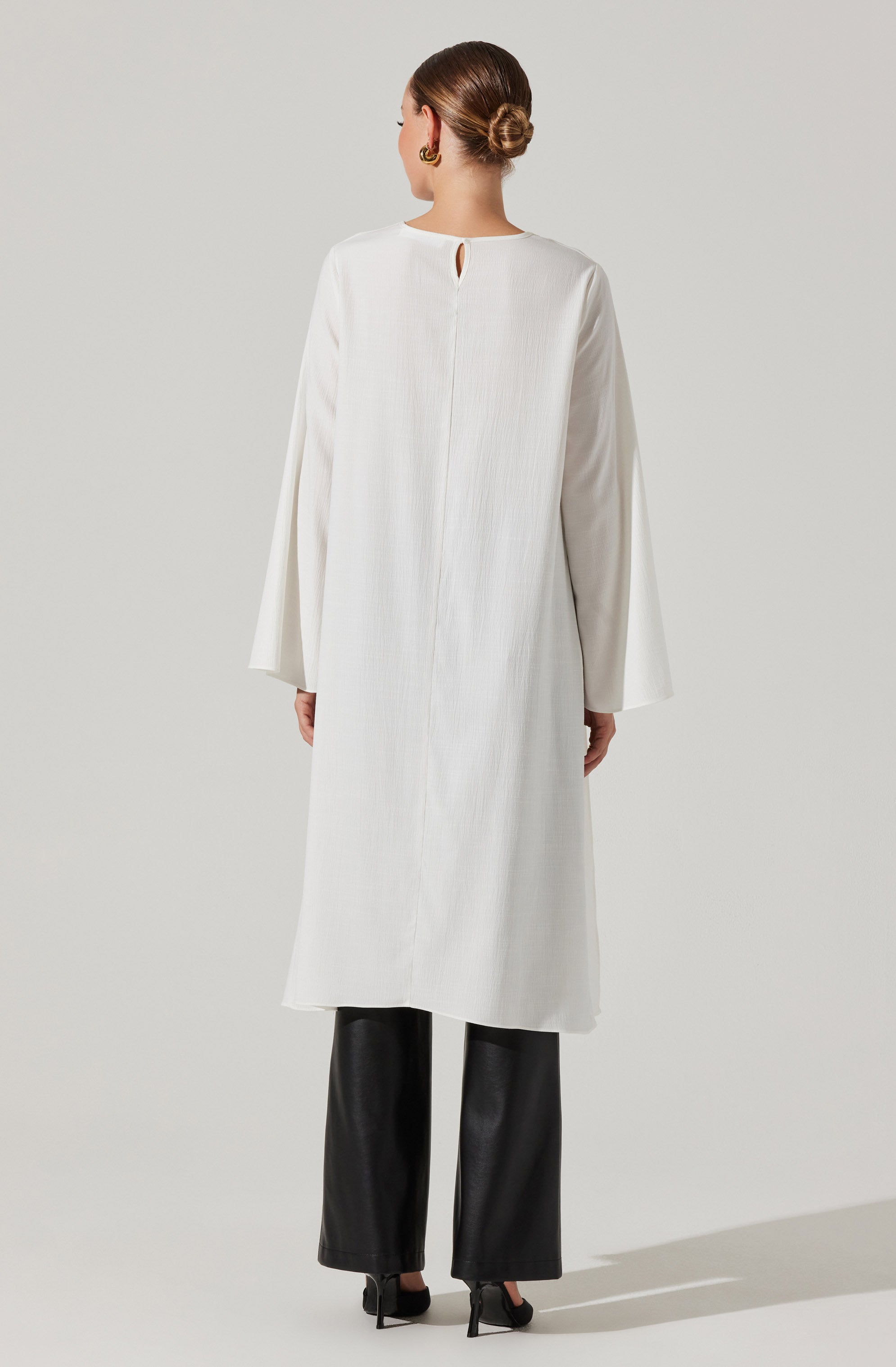 A woman is wearing a long, white, textured coat with a small keyhole opening at the back neckline. The coat has long, wide sleeves and reaches below the knees. She is also wearing black wide-leg pants and black high-heeled shoes.