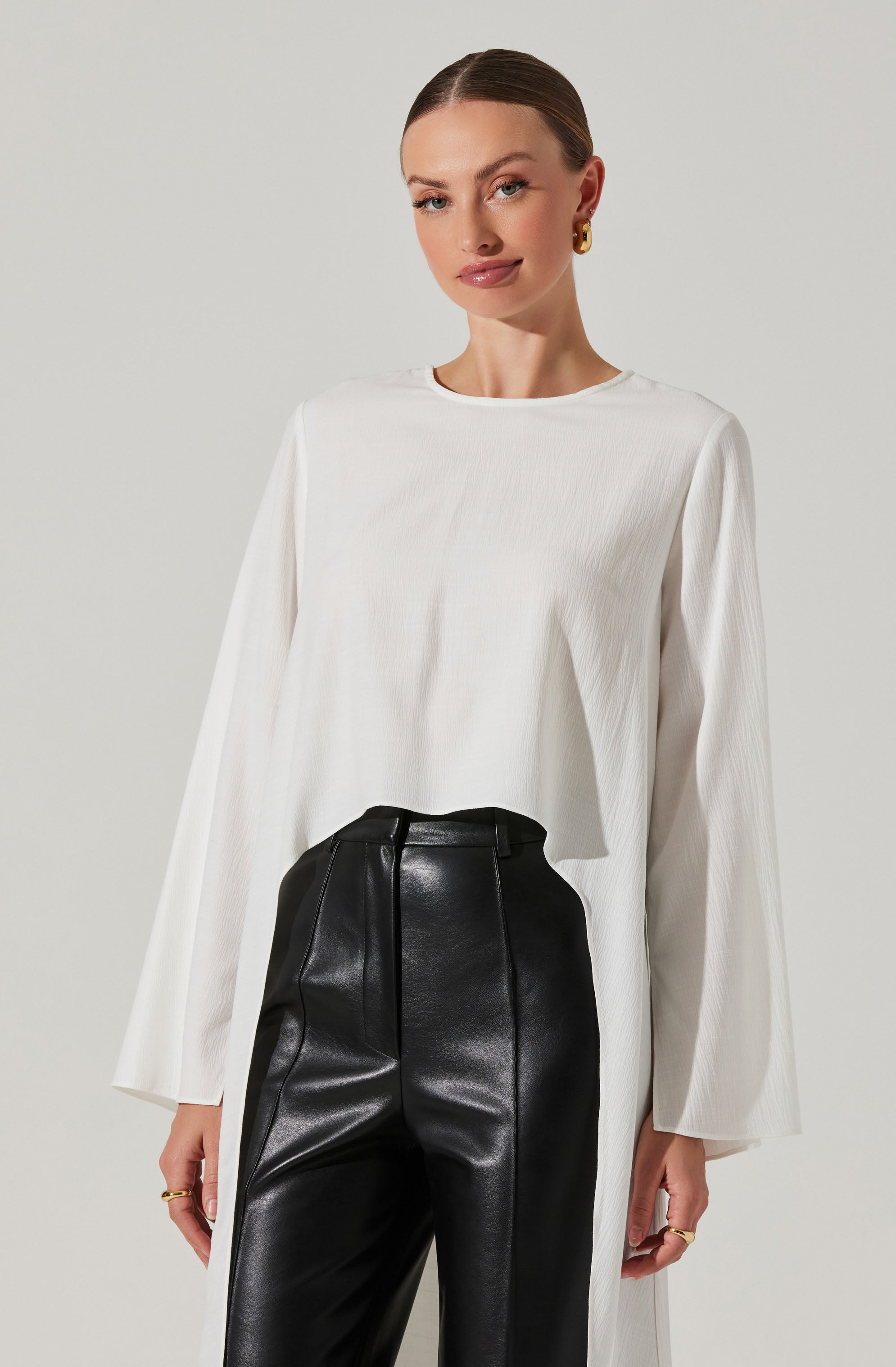 A woman is wearing a loose-fitting, long-sleeved white top made of a lightweight, slightly textured fabric. She pairs it with high-waisted black leather pants that have a smooth finish and prominent vertical stitching down the front.