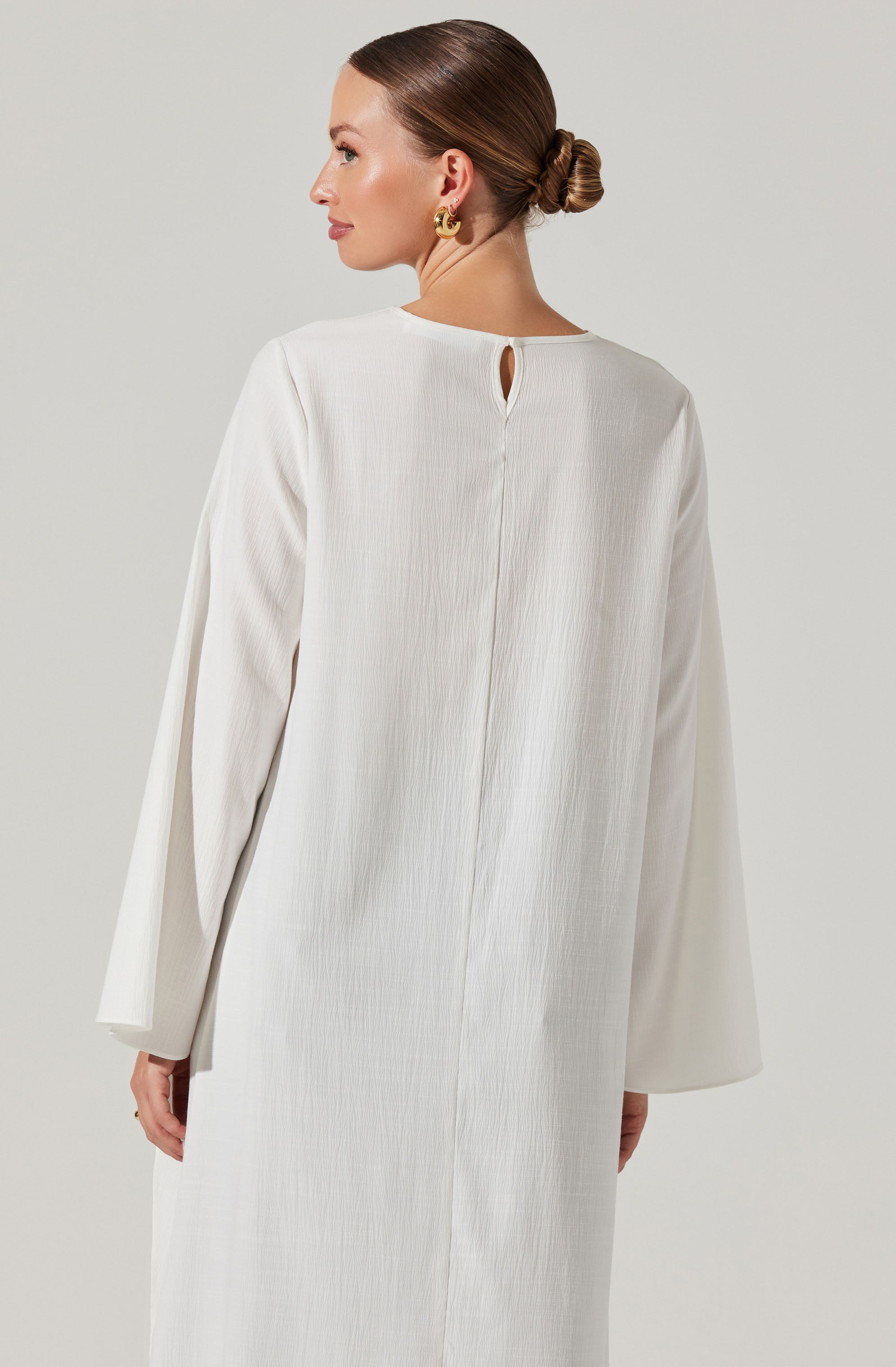 The image shows a woman wearing a long, white, textured dress with long sleeves. The dress has a small keyhole opening at the back near the neckline. The fabric appears to be lightweight and slightly crinkled. The woman has her hair styled in a low bun and is accessorized with gold earrings.