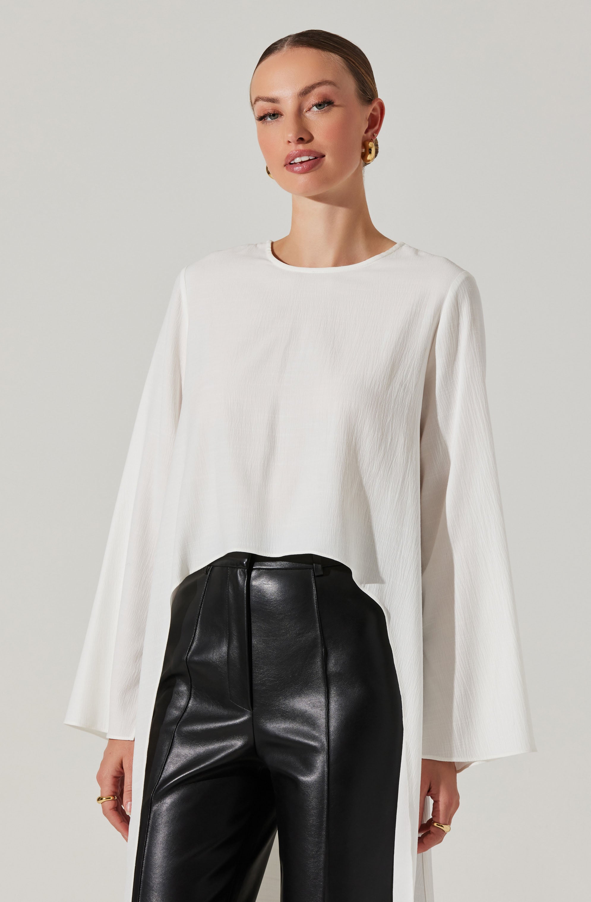 A woman is wearing a loose-fitting, long-sleeved white top with a round neckline and wide sleeves. She pairs it with high-waisted black leather pants that have a smooth, shiny finish. She accessorizes with large gold hoop earrings and rings on her fingers.