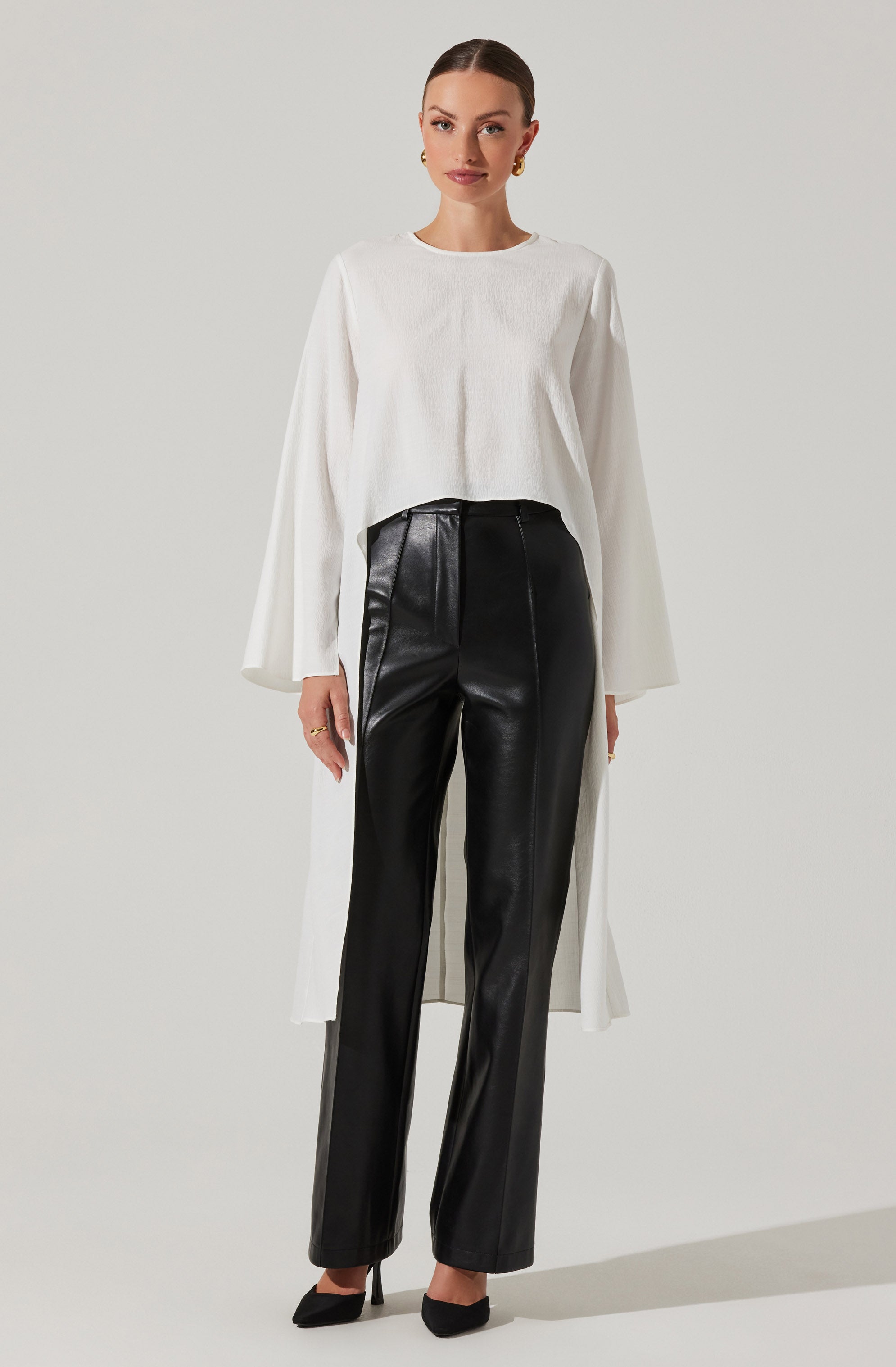 The woman is wearing a loose-fitting, long-sleeved white top with a round neckline. She has paired it with black, high-waisted leather pants that have a straight-leg cut. She is also wearing black pointed-toe high heels.