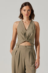 A woman is wearing a sleeveless, olive-green top with a deep V-neckline that is knotted at the waist, paired with matching high-waisted, pleated trousers that have a belt loop and a single button closure. The overall style is sophisticated and modern.