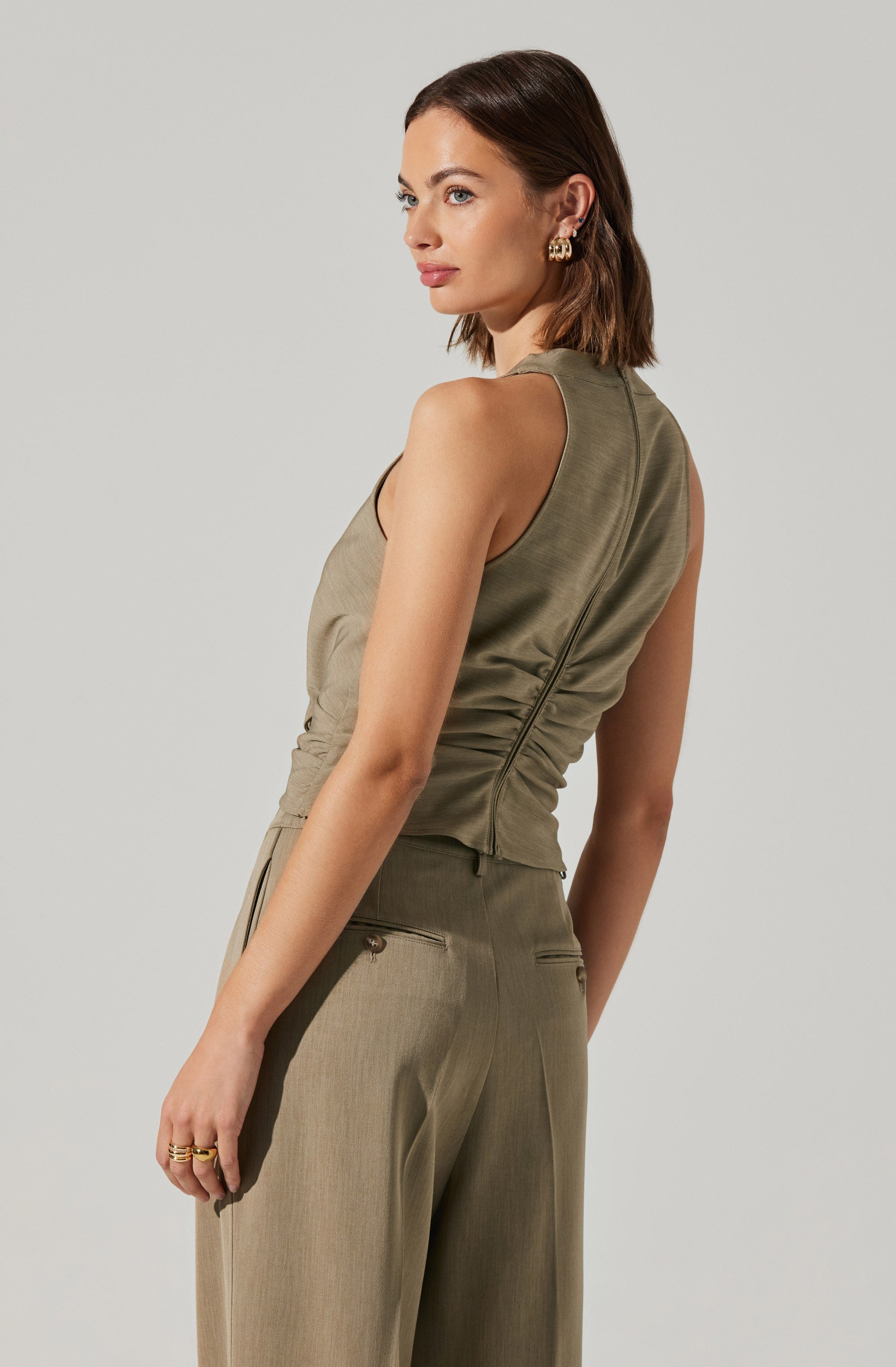 A woman is wearing a sleeveless, olive-green top with a high neckline and visible side seam detail. She is also wearing matching loose-fitting, beige trousers with zippered back pockets.