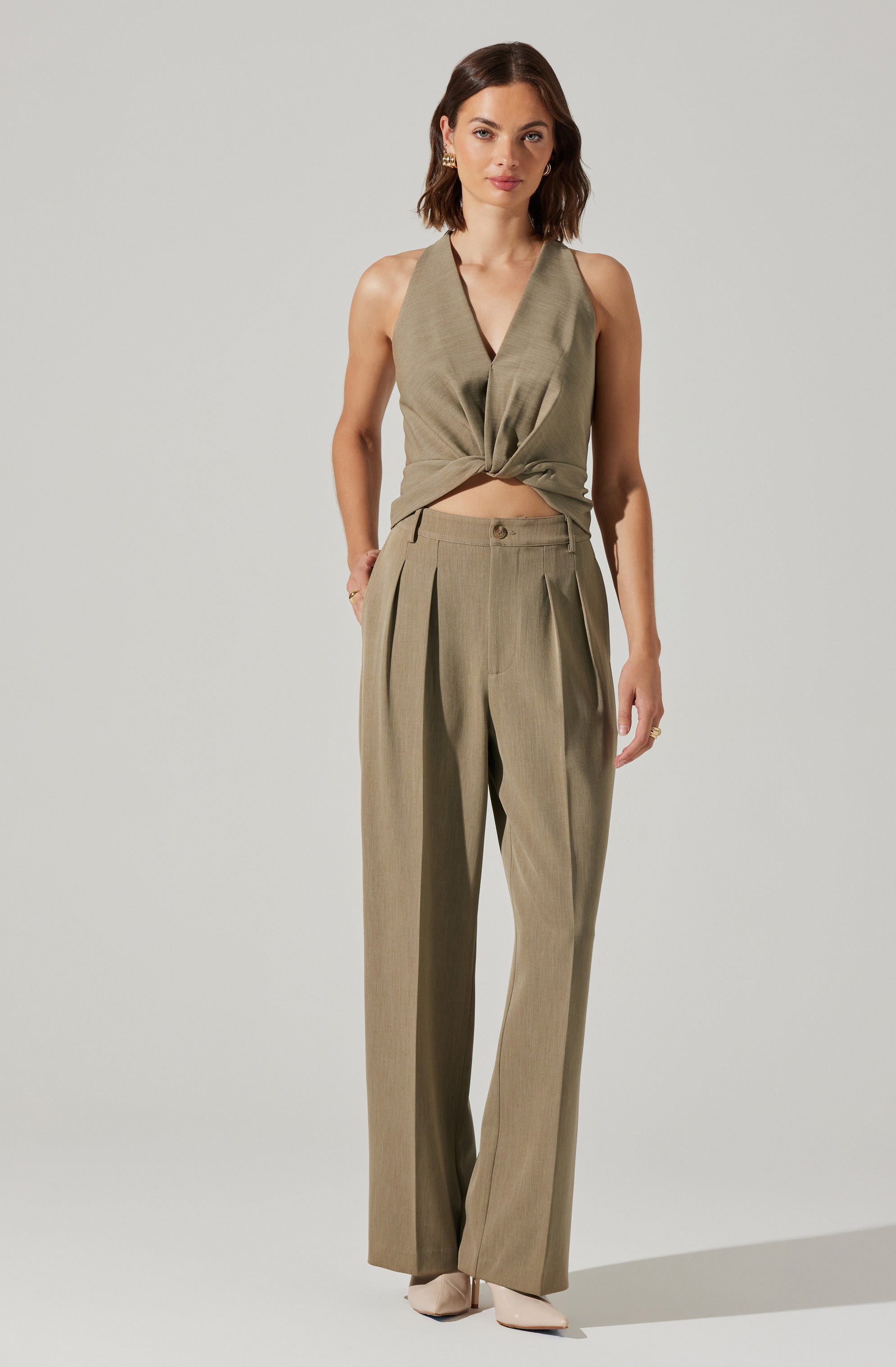 A woman is wearing a sleeveless, olive-green, satin-like top with a deep V neckline that is tied in a knot at the waist. She is also dressed in high-waisted, wide-leg trousers in a matching olive-green color, featuring pleats at the front and a button closure. She has on light-colored shoes and is accessorized with earrings and a ring.