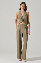 A woman is wearing a sleeveless, olive-green, satin-like top with a deep V neckline that is tied in a knot at the waist. She is also dressed in high-waisted, wide-leg trousers in a matching olive-green color, featuring pleats at the front and a button closure. She has on light-colored shoes and is accessorized with earrings and a ring.