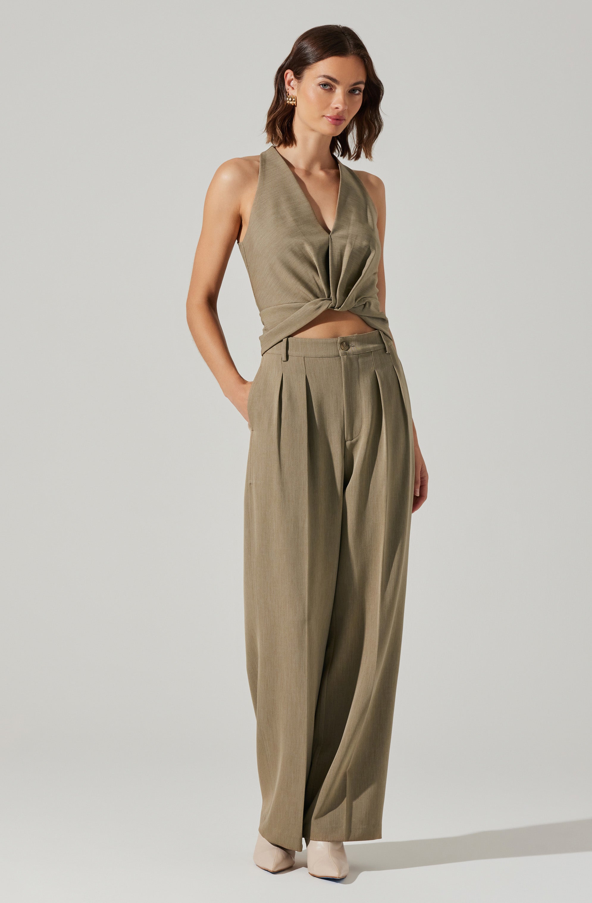 A woman is wearing a sleeveless, olive-green top with a v-neckline and a front knot detail. She pairs it with high-waisted, wide-leg matching olive-green trousers that feature pleats at the waist. She also has on light-colored shoes.