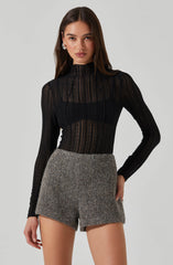 A woman is wearing a black, long-sleeved, semi-sheer, high-neck top with vertical striped and lace-like pattern details. She pairs it with high-waisted, textured gray shorts. She also has on statement earrings.