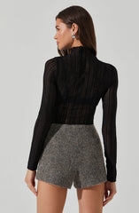 A woman is wearing a fitted black long-sleeve top with a high neckline made of a sheer, ribbed, and slightly textured fabric. She pairs it with high-waisted, gray tweed shorts that have a subtle speckled pattern. She has long, straight brown hair and is wearing statement silver earrings.