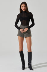 A woman is wearing a sheer black, long-sleeved, high-neck top with vertical stripe patterns. She pairs it with high-waisted, gray, textured shorts. She completes her outfit with tall, black leather knee-high boots with high heels.