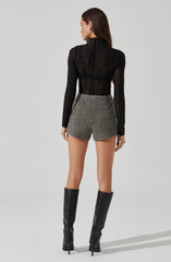 The woman is wearing a black, long-sleeved, semi-sheer, striped top with a high neckline. She has paired it with high-waisted, gray, textured shorts. She is also wearing tall black leather boots that reach above the knees. Her hair is long and brown, and she is accessorized with earrings.