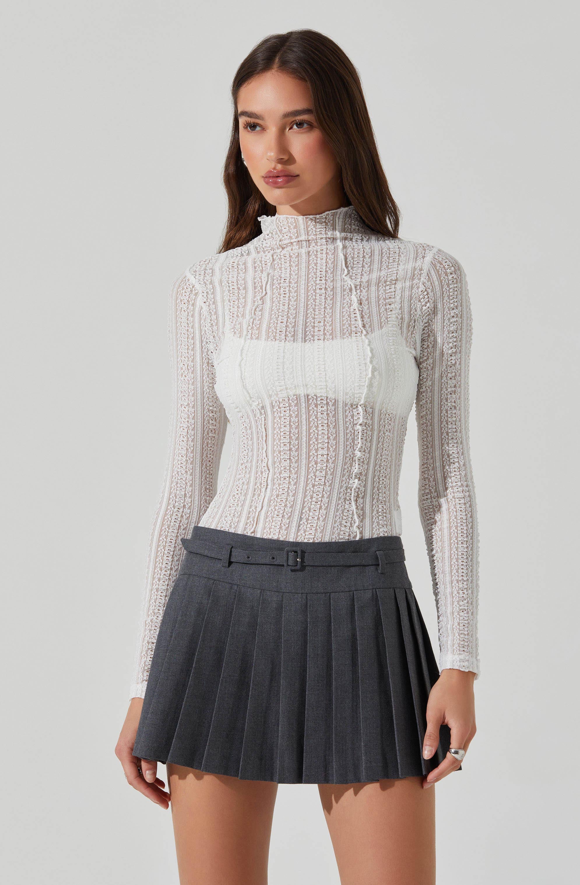 A woman is wearing a high-neck, long-sleeved, sheer white top with a textured, lace-like pattern. She pairs it with a dark gray, pleated mini skirt that features a waistband with a small buckle and belt loops.