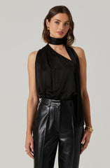 A woman is wearing a sleeveless, satin black top with a draped design and a matching choker collar. She pairs it with high-waisted, black leather trousers that have a pleated front and a slightly loose fit. She accessorizes with gold earrings, a gold bracelet, and a gold ring.