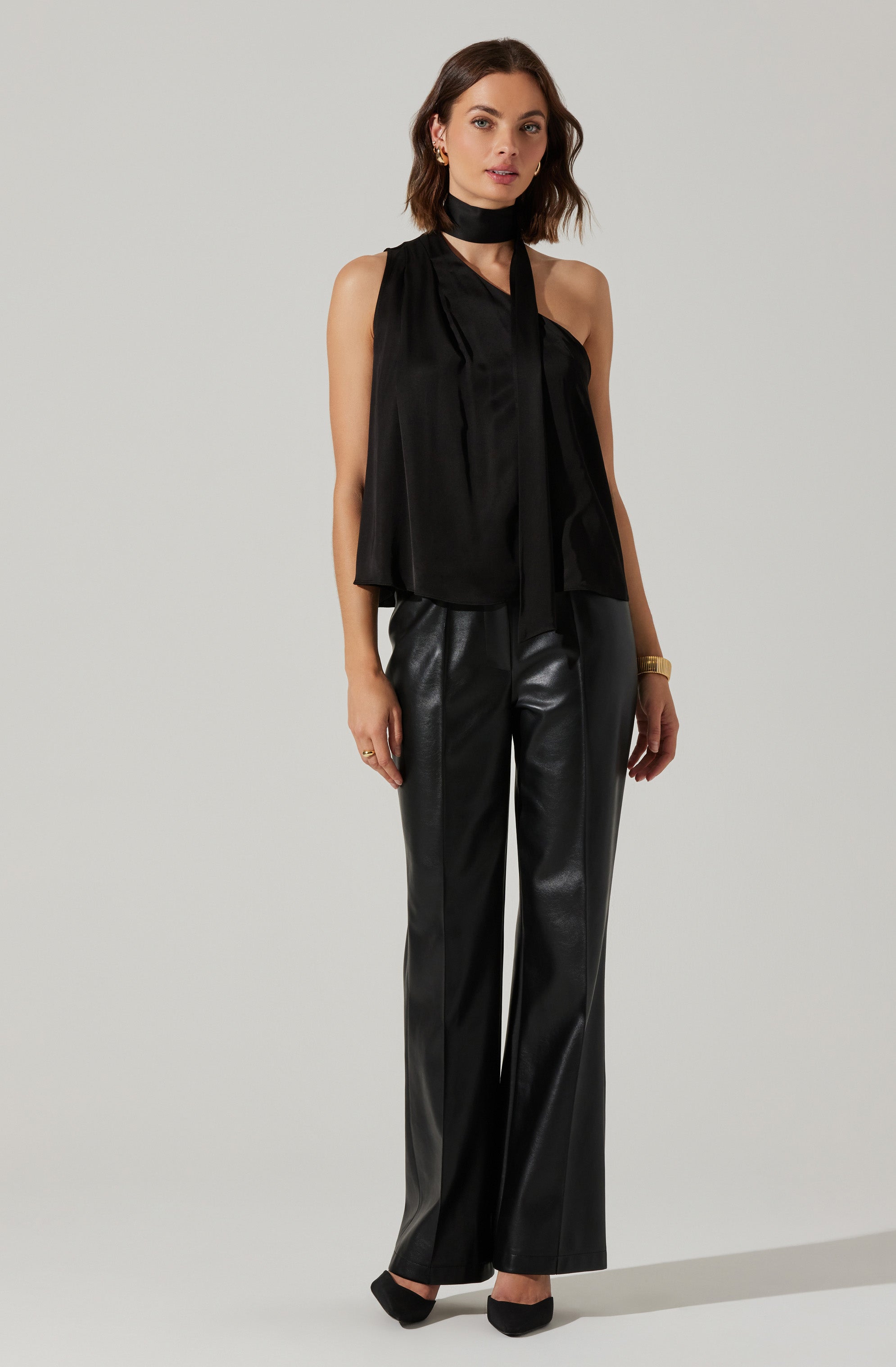 A woman is wearing a sleeveless black top with a high neck and a long, narrow tie at the neckline. She pairs it with high-waisted, wide-leg black leather pants. She accessorizes with small earrings, a gold bracelet, and black high heels.