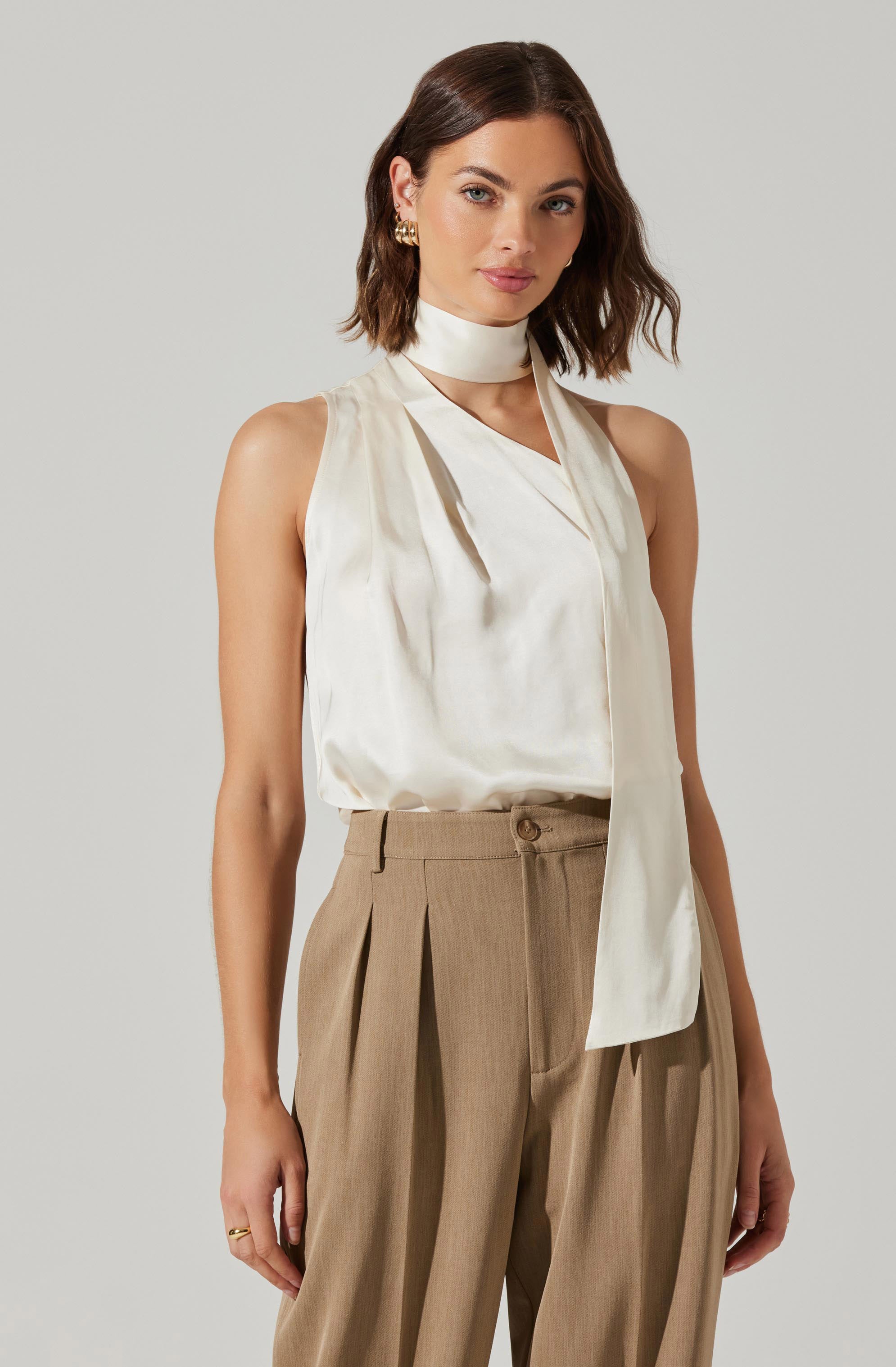 A woman is wearing a silky, sleeveless, off-white top with a wrap detail and a high neck that extends into a long, narrow tie. She is also wearing high-waisted, wide-leg beige trousers with pleats at the front.